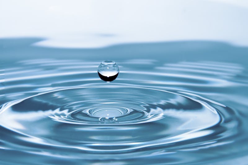 Single clear water droplet suspended above concentric ripples on a calm blue water surface